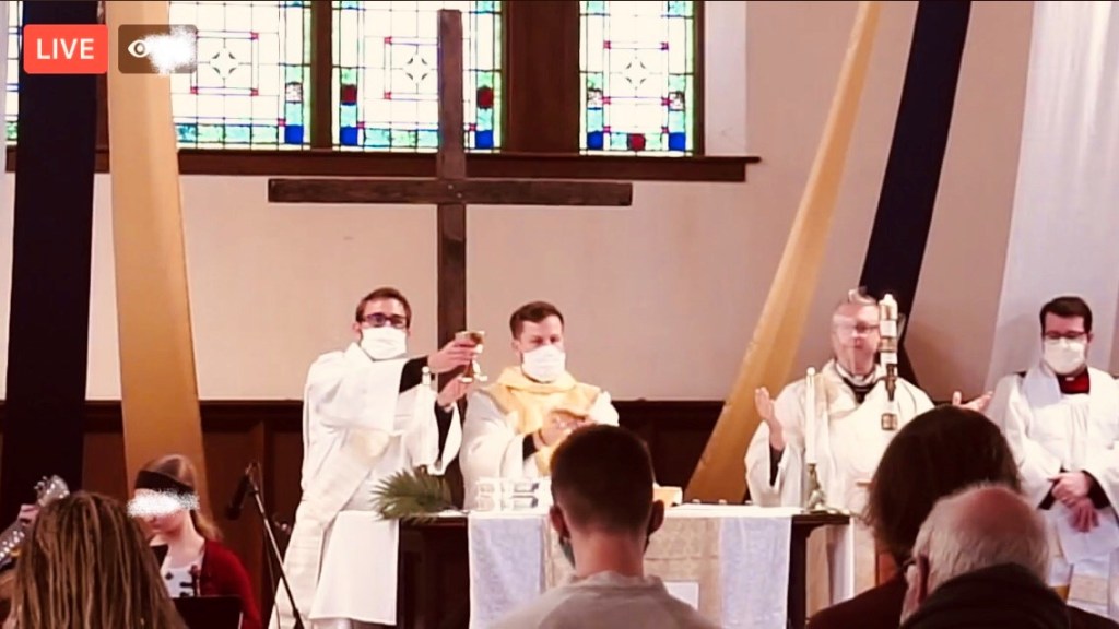 At the sacramental table, deacon and priest hold up the cup and bread in a front of stained glass windows