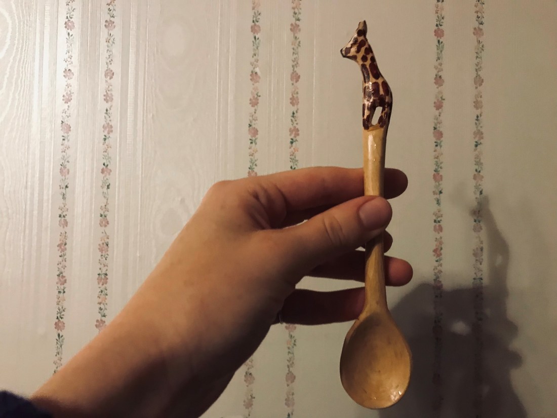 A hand holds a small wooden spoon designed with a giraffe on the handle. The backdrop is a flowery white wall.