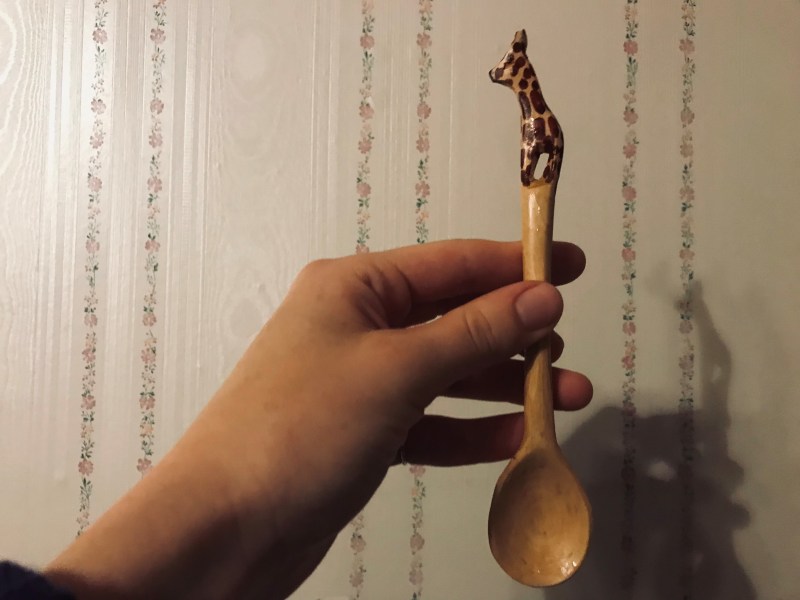 A hand holds a small wooden spoon designed with a giraffe on the handle. The backdrop is a flowery white wall.